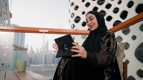 احتساء المشروب المفضل في الكافيه، ادارة الأعمال وانجاز المهام المطلوبة، العمل عن بعد، مهنة ووظيفة نسائية، تصوير مقرب لإمرأة عربية خليجية اماراتية محجبة ترتدي عباءة ملونة تستخدم الجهاز اللوحي وتشرب القهوة، استخدام جهاز تقني حديث ومتطور