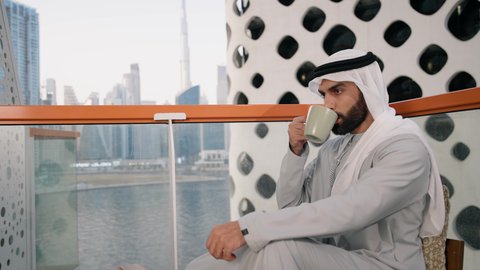 الاستمتاع بالمشروبات الساخنة، تصوير مقرب لرجل عربي خليجي اماراتي يرتدي الكندورة والغترة البيضاء يجلس على اريكة مريحة يقوم بشرب القهوة في المقهى، قضاء وقت الفراغ باحتساء المشروب المفضل، تناول الأعشاب الشتوية الدافئة