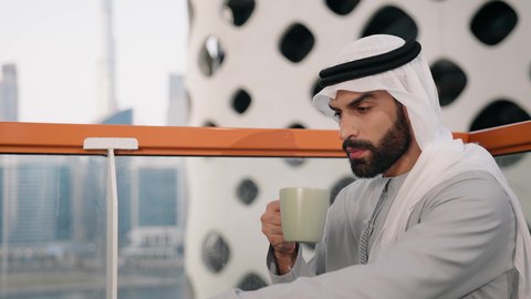 قضاء وقت الفراغ باحتساء المشروب المفضل، تناول الأعشاب الشتوية الدافئة، الاستمتاع بالمشروبات الساخنة، تصوير مقرب لرجل عربي خليجي اماراتي يرتدي الكندورة والغترة البيضاء يجلس على اريكة مريحة يقوم بشرب القهوة في المقهى