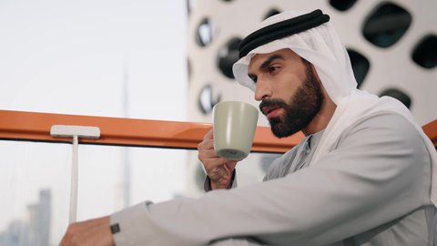 الاستمتاع بالمشروبات الساخنة، تصوير مقرب لرجل عربي خليجي اماراتي يرتدي الكندورة والغترة البيضاء يجلس على اريكة مريحة يقوم بشرب القهوة في المقهى، قضاء وقت الفراغ باحتساء المشروب المفضل، تناول الأعشاب الشتوية الدافئة