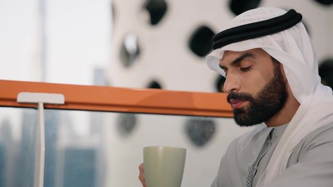 تناول الأعشاب الشتوية الدافئة، الاستمتاع بالمشروبات الساخنة، تصوير مقرب لرجل عربي خليجي اماراتي يرتدي الكندورة والغترة البيضاء يجلس على اريكة مريحة يقوم بشرب القهوة في المقهى، قضاء وقت الفراغ باحتساء المشروب المفضل