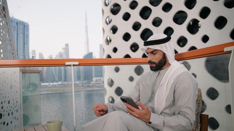 Sipping your favorite drink, a close-up of an Emirati Gulf Arab man wearing a kandura and a white ghutra holding a mobile phone and a cup of coffee, using a modern and advanced technical device, working remotely, communicating and exchanging conversations with family and friends, browsing social media applications and programs