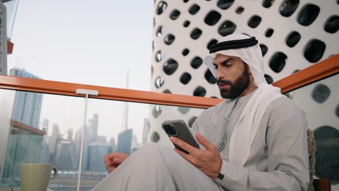 العمل عن بعد، استخدام جهاز تقني حديث ومتطور،  تبادل الأحاديث مع الأهل والأصدقاء، تصفح التطبيقات وبرامج التواصل الاجتماعي، احتساء المشروب المفضل، تصوير مقرب لرجل عربي خليجي اماراتي يرتدي الكندورة والغترة البيضاء يمسك بيده الهاتف المحمول وكوب من القهوة