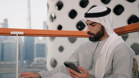 Using a modern and advanced technical device, working remotely, exchanging conversations with family and friends, browsing applications and social networking programs, sipping a favorite drink, a close-up photo of an Emirati Gulf Arab man wearing a kandura and a white ghutra, holding a mobile phone and a cup of coffee.