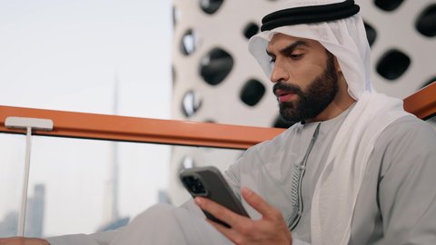 احتساء المشروب المفضل، تصوير مقرب لرجل عربي خليجي اماراتي يرتدي الكندورة والغترة البيضاء يمسك بيده الهاتف المحمول وكوب من القهوة، استخدام جهاز تقني حديث ومتطور، العمل عن بعد،  تبادل الأحاديث مع الأهل والأصدقاء، تصفح التطبيقات وبرامج التواصل الاجتماعي