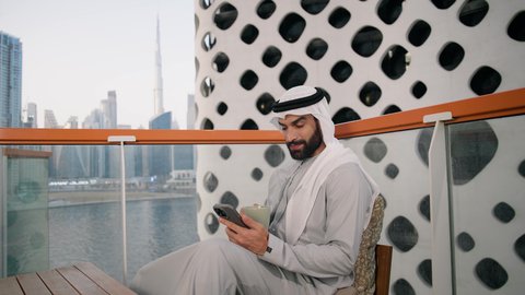 Exchanging conversations, communication and news with family and friends, a close-up of an Emirati Gulf Arab man wearing a kandura and a white ghutra holding a mobile phone, using a modern and advanced technical device, working remotely, browsing applications and social networking programs
