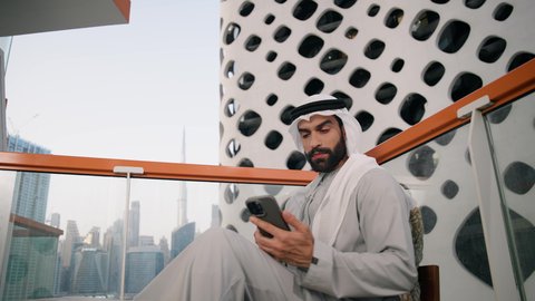 التواصل و تبادل الأحاديث والأخبار مع الأهل والأصدقاء، تصوير مقرب لرجل عربي خليجي اماراتي يرتدي الكندورة والغترة البيضاء يمسك بيده الهاتف المحمول، استخدام جهاز تقني حديث ومتطور، العمل عن بعد، تصفح التطبيقات وبرامج التواصل الاجتماعي