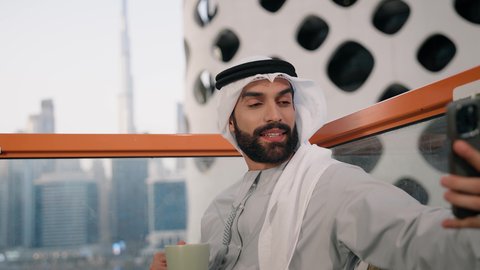 توثيق اللحظات السعيدة، التقاط الصور الشخصية باستخدام الجوال، استخدام جهاز تقني حديث ومتطور في تخليد الذكريات، تصوير مقرب لرجل عربي خليجي اماراتي يرتدي الكندورة والغترة البيضاء يمسك الهاتف المحمول ويأخذ صورة سيلفي بإيماءات السرور