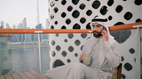 Making a phone call, using a modern and advanced technical device, sipping your favorite drink, a close-up of an Emirati Gulf Arab man wearing a white kandura and ghutra holding a mobile phone and a cup of coffee, working remotely, communicating and exchanging conversations with family and friends