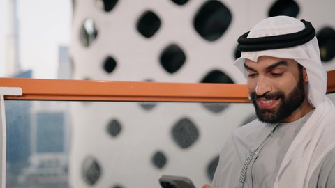 تبادل الأحاديث والتواصل والأخبار مع الأهل والأصدقاء، تصوير مقرب لرجل عربي خليجي اماراتي يرتدي الكندورة والغترة البيضاء يمسك بيده الهاتف المحمول، استخدام جهاز تقني حديث ومتطور، العمل عن بعد، تصفح التطبيقات وبرامج التواصل الاجتماعي