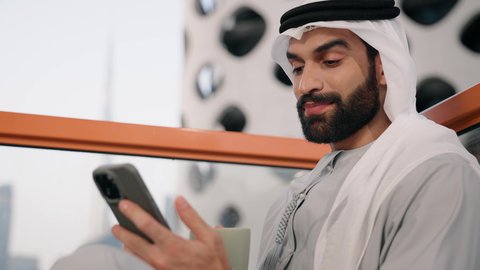 التواصل و تبادل الأحاديث والأخبار مع الأهل والأصدقاء، تصوير مقرب لرجل عربي خليجي اماراتي يرتدي الكندورة والغترة البيضاء يمسك بيده الهاتف المحمول، استخدام جهاز تقني حديث ومتطور، العمل عن بعد، تصفح التطبيقات وبرامج التواصل الاجتماعي