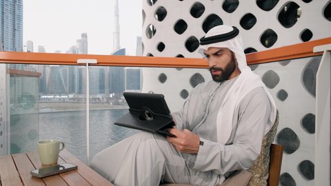 أداء مهام العمل عن بعد، إدارة شؤون الأعمال وتنظيمها عبر الايباد، ايماءات السرور والسعادة، استخدام جهاز تقني حديث ومتطور، تصوير مقرب لرجل عربي خليجي اماراتي يرتدي الكندورة والغترة البيضاء يستخدم الجهاز اللوحي