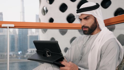 إدارة شؤون الأعمال وتنظيمها عبر الايباد، ايماءات السرور والسعادة، استخدام جهاز تقني حديث ومتطور، تصوير مقرب لرجل عربي خليجي اماراتي يرتدي الكندورة والغترة البيضاء يستخدم الجهاز اللوحي، أداء مهام العمل عن بعد