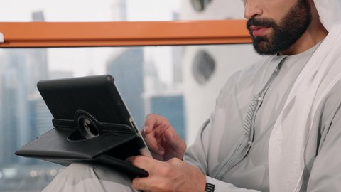تنظيم شؤون الأعمال وادارتها عبر الايباد، ايماءات السرور والسعادة، استخدام جهاز تقني حديث ومتطور، تصوير مقرب لرجل عربي خليجي اماراتي يرتدي الكندورة والغترة البيضاء يستخدم الجهاز اللوحي، أداء مهام العمل عن بعد