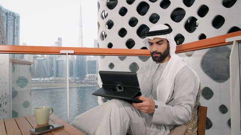 تنظيم شؤون الأعمال وادارتها عبر الايباد، ايماءات السرور والسعادة، استخدام جهاز تقني حديث ومتطور، تصوير مقرب لرجل عربي خليجي اماراتي يرتدي الكندورة والغترة البيضاء يستخدم الجهاز اللوحي، أداء مهام العمل عن بعد