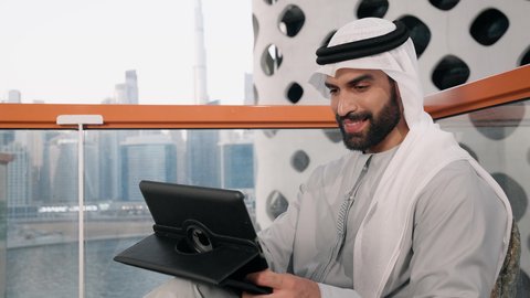 إدارة شؤون الأعمال وتنظيمها عبر الايباد، ايماءات السرور والسعادة، استخدام جهاز تقني حديث ومتطور، تصوير مقرب لرجل عربي خليجي اماراتي يرتدي الكندورة والغترة البيضاء يستخدم الجهاز اللوحي، أداء مهام العمل عن بعد