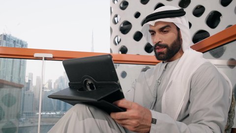 تنظيم شؤون الأعمال وادارتها عبر الايباد، ايماءات السرور والسعادة، استخدام جهاز تقني حديث ومتطور، تصوير مقرب لرجل عربي خليجي اماراتي يرتدي الكندورة والغترة البيضاء يستخدم الجهاز اللوحي، أداء مهام العمل عن بعد