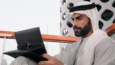 ايماءات السرور والسعادة، استخدام جهاز تقني حديث ومتطور، تصوير مقرب لرجل عربي خليجي اماراتي يرتدي الكندورة والغترة البيضاء يستخدم الجهاز اللوحي، إنجاز مهام العمل عن بعد، إدارة شؤون الأعمال وتنظيمها عبر الايباد
