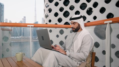 استخدام جهاز تقني حديث ومتطور، تصوير مقرب لرجل عربي خليجي اماراتي يرتدي الكندورة والغترة البيضاء يستخدم الحاسوب المحمول، إنجاز مهام العمل عن بعد، إدارة شؤون الأعمال وتنظيمها عبر اللابتوب، ايماءات السعادة والسرور