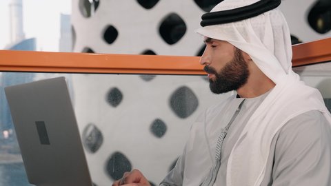استخدام جهاز تقني حديث ومتطور، تصوير مقرب لرجل عربي خليجي اماراتي يرتدي الكندورة والغترة البيضاء يستخدم الحاسوب المحمول، إنجاز مهام العمل عن بعد، إدارة شؤون الأعمال وتنظيمها عبر اللابتوب، ايماءات السعادة والسرور