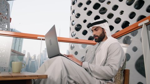 Using a modern and advanced technical device, a close-up of an Emirati Gulf Arab man wearing a white kandura and ghutra using a laptop, completing work tasks remotely, managing and organizing business affairs via the laptop, gestures of happiness and pleasure