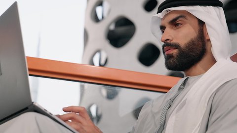 إتمام مهام العمل عن بعد، إدارة شؤون الأعمال وتنظيمها عبر اللابتوب، ايماءات السعادة والسرور، استخدام جهاز تقني حديث ومتطور، تصوير مقرب لرجل عربي خليجي اماراتي يرتدي الكندورة والغترة البيضاء يستخدم الحاسوب المحمول