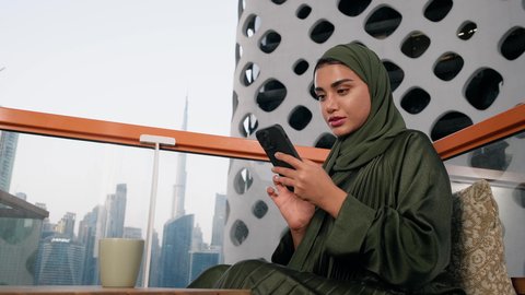 Sipping your favorite drink at a cafe, browsing social media apps and programs, exchanging conversations and news with family and friends, a close-up of an Emirati woman wearing an abaya and hijab holding a mobile phone and drinking coffee, using a modern and advanced technical device
