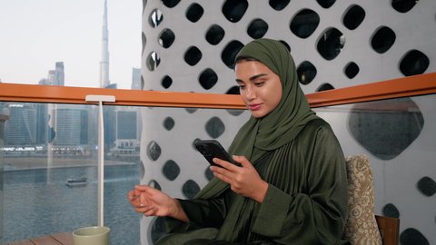 تصفح التطبيقات وبرامج التواصل الاجتماعي، تبادل الأحاديث والأخبار مع الأهل والأصدقاء،  تصوير مقرب لامراة اماراتية ترتدي العباءة والحجاب تمسك بيدها الهاتف المحمول وتشرب القهوة، استخدام جهاز تقني حديث ومتطور، احتساء المشروب المفضل في المقهى