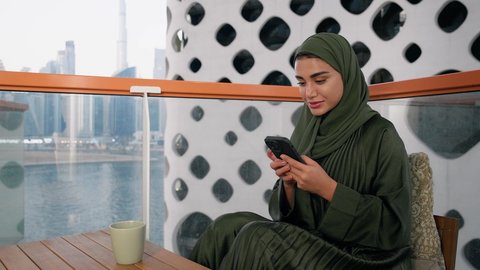 Browsing social media apps and programs, exchanging conversations and news with family and friends, a close-up of an Emirati woman wearing an abaya and hijab holding a mobile phone and drinking coffee, using a modern and advanced technical device, sipping a favorite drink in a café