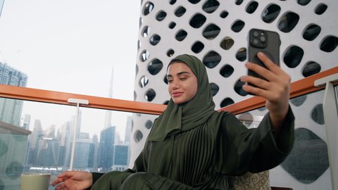 التقاط الصور الشخصية باستخدام الهاتف المحمول، تصوير مقرب لامراة اماراتية ترتدي العباءة والحجاب تأخذ صورة سيلفي بإيماءات السعادة والسرور، توثيق اللحظات التذكارية السعيدة، استخدام جهاز تقني حديث ومتطور لتخليد الذكريات