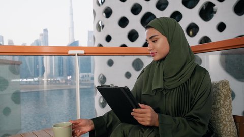 شرب المشروب المفضل في الكافيه، ادارة الأعمال وانجاز المهام المطلوبة، العمل عن بعد، مهنة ووظيفة نسائية، تصوير مقرب لإمرأة عربية خليجية اماراتية محجبة ترتدي عباءة ملونة تستخدم الجهاز اللوحي وتشرب القهوة، استخدام جهاز تقني حديث ومتطور