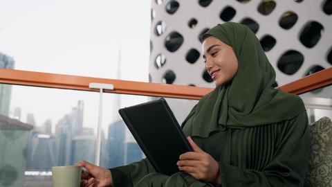 شرب المشروب المفضل في الكافيه، ادارة الأعمال وانجاز المهام المطلوبة، العمل عن بعد، مهنة ووظيفة نسائية، تصوير مقرب لإمرأة عربية خليجية اماراتية محجبة ترتدي عباءة ملونة تستخدم الجهاز اللوحي وتشرب القهوة، استخدام جهاز تقني حديث ومتطور