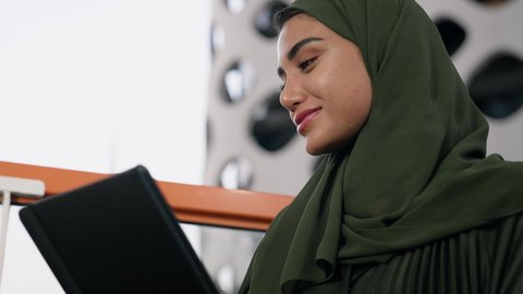 Sipping your favorite drink at a cafe, managing your business and completing your tasks, working remotely, a female profession and job, a close-up of a veiled Emirati Gulf Arab woman wearing a colorful abaya using a tablet and drinking coffee, using a modern and advanced technical device