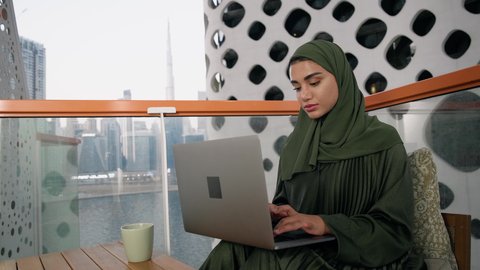 ايماءات التركيز والاندماج، ادارة الأعمال وانجاز المهام المطلوبة، العمل عن بعد،  مهنة ووظيفة نسائية،  تصوير مقرب لإمرأة عربية خليجية اماراتية محجبة ترتدي عباءة ملونة تستخدم الحاسوب المحمول، استخدام جهاز تقني حديث ومتطور