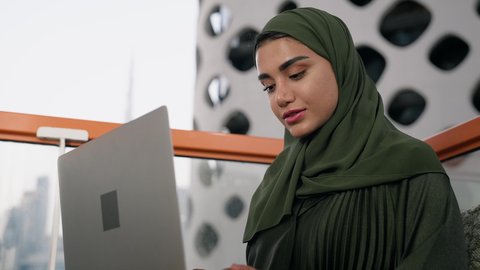 وظيفة ومهنة نسائية،  تصوير مقرب لإمرأة عربية خليجية اماراتية محجبة ترتدي عباءة ملونة تستخدم الحاسوب المحمول، استخدام جهاز تقني حديث ومتطور، ايماءات التركيز والاندماج، ادارة الأعمال وانجاز المهام المطلوبة، العمل عن بعد