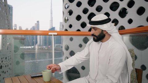 الاستمتاع بالمشروبات الساخنة، تصوير مقرب لرجل عربي خليجي اماراتي يرتدي الكندورة والغترة البيضاء يجلس على اريكة مريحة يقوم بشرب القهوة في المقهى، إيماءات السعادة والسرور، قضاء وقت الفراغ باحتساء المشروب المفضل، تناول الأعشاب الشتوية الدافئة