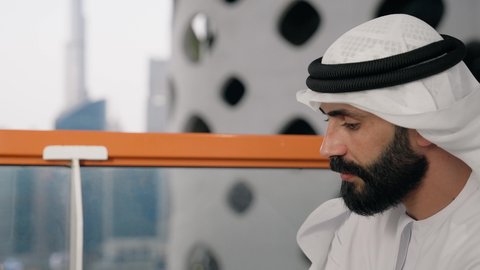 قضاء وقت الفراغ باحتساء المشروب المفضل, تناول الأعشاب الشتوية الدافئة، الاستمتاع بالمشروبات الساخنة، تصوير مقرب لرجل عربي خليجي اماراتي يرتدي الكندورة والغترة البيضاء يجلس على اريكة مريحة يقوم بشرب القهوة في المقهى، إيماءات السعادة والسرور