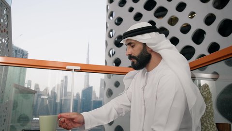 الاستمتاع بالمشروبات الساخنة، تصوير مقرب لرجل عربي خليجي اماراتي يرتدي الكندورة والغترة البيضاء يجلس على اريكة مريحة يقوم بشرب القهوة في المقهى، إيماءات السعادة والسرور، قضاء وقت الفراغ باحتساء المشروب المفضل، تناول الأعشاب الشتوية الدافئة