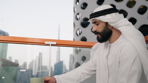 تناول الأعشاب الشتوية الدافئة، الاستمتاع بالمشروبات الساخنة، تصوير مقرب لرجل عربي خليجي اماراتي يرتدي الكندورة والغترة البيضاء يجلس على اريكة مريحة يقوم بشرب القهوة في المقهى، إيماءات السعادة والسرور، قضاء وقت الفراغ باحتساء المشروب المفضل