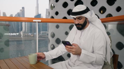 Working remotely, browsing social media apps and programs, exchanging conversations and news with family and friends, a close-up of an Emirati Gulf Arab man wearing a white kandura and ghutra holding a mobile phone, using a modern and advanced technical device