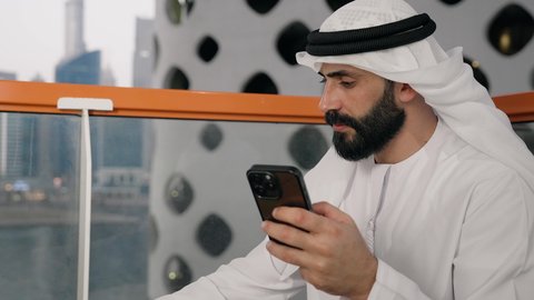 تبادل الأحاديث والأخبار مع الأهل والأصدقاء، تصوير مقرب لرجل عربي خليجي اماراتي يرتدي الكندورة والغترة البيضاء يمسك بيده الهاتف المحمول، استخدام جهاز تقني حديث ومتطور، العمل عن بعد، تصفح التطبيقات وبرامج التواصل الاجتماعي