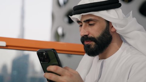 تصفح التطبيقات وبرامج التواصل الاجتماعي، تبادل الأحاديث والأخبار مع الأهل والأصدقاء، تصوير مقرب لرجل عربي خليجي اماراتي يرتدي الكندورة والغترة البيضاء يمسك بيده الهاتف المحمول، استخدام جهاز تقني حديث ومتطور، العمل عن بعد