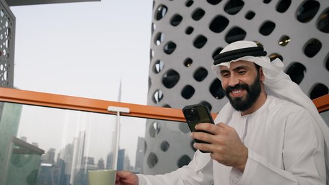 التواصل و تبادل الأحاديث والأخبار مع الأهل والأصدقاء، تصوير مقرب لرجل عربي خليجي اماراتي يرتدي الكندورة والغترة البيضاء يمسك بيده الهاتف المحمول، استخدام جهاز تقني حديث ومتطور، العمل عن بعد، تصفح التطبيقات وبرامج التواصل الاجتماعي
