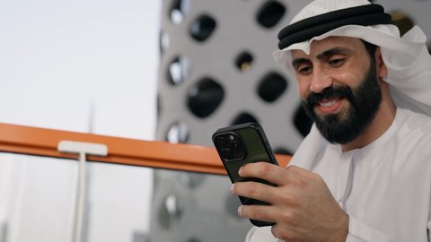 التواصل و تبادل الأحاديث والأخبار مع الأهل والأصدقاء، تصوير مقرب لرجل عربي خليجي اماراتي يرتدي الكندورة والغترة البيضاء يمسك بيده الهاتف المحمول، استخدام جهاز تقني حديث ومتطور، العمل عن بعد، تصفح التطبيقات وبرامج التواصل الاجتماعي