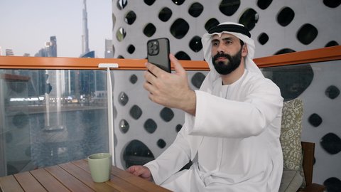 Taking selfies with a mobile phone, using a modern and advanced technical device to immortalize memories, a close-up of an Emirati Gulf Arab man wearing a kandura and a white ghutra holding a mobile phone and taking a selfie with gestures of pleasure, documenting happy moments