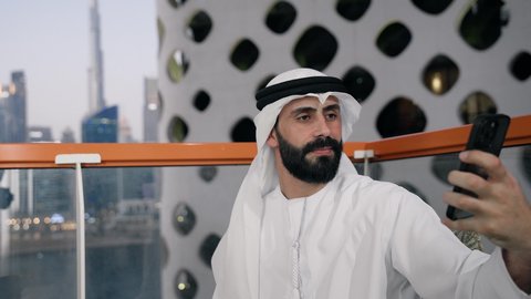 Documenting happy moments, taking selfies with a mobile phone, using a modern and advanced technical device to immortalize memories, a close-up of an Emirati Gulf Arab man wearing a kandura and a white ghutra holding a mobile phone and taking a selfie with gestures of pleasure