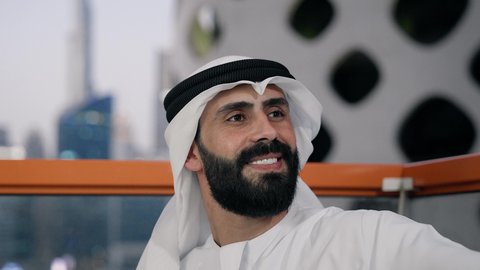 التقاط الصور الشخصية باستخدام الجوال، استخدام جهاز تقني حديث ومتطور في تخليد الذكريات، تصوير مقرب لرجل عربي خليجي اماراتي يرتدي الكندورة والغترة البيضاء يمسك الهاتف المحمول ويأخذ صورة سيلفي بإيماءات السرور، توثيق اللحظات السعيدة