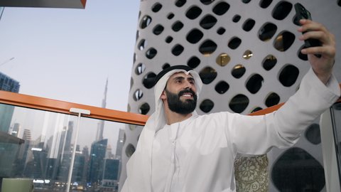 تصوير مقرب لرجل عربي خليجي اماراتي يرتدي الكندورة والغترة البيضاء يمسك الهاتف المحمول ويأخذ صورة سيلفي بإيماءات السرور، توثيق اللحظات السعيدة، التقاط الصور الشخصية باستخدام الجوال، استخدام جهاز تقني حديث ومتطور في تخليد الذكريات