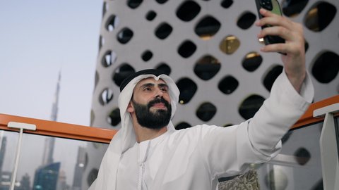 تصوير مقرب لرجل عربي خليجي اماراتي يرتدي الكندورة والغترة البيضاء يمسك الهاتف المحمول ويأخذ صورة سيلفي بإيماءات السرور، توثيق اللحظات السعيدة، التقاط الصور الشخصية باستخدام الجوال، استخدام جهاز تقني حديث ومتطور في تخليد الذكريات