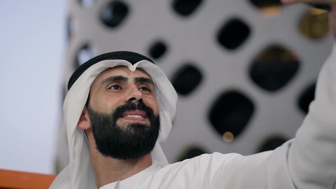 تصوير مقرب لرجل عربي خليجي اماراتي يرتدي الكندورة والغترة البيضاء يمسك الهاتف المحمول ويأخذ صورة سيلفي بإيماءات السرور، توثيق اللحظات السعيدة، التقاط الصور الشخصية باستخدام الجوال، استخدام جهاز تقني حديث ومتطور في تخليد الذكريات
